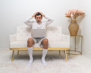 A man sits on a modern white couch, holding his head in surprise while interacting with his laptop in a minimalist living room.