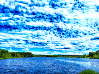 A vibrant blue sky filled with fluffy clouds reflects over a calm lake bordered by lush greenery.