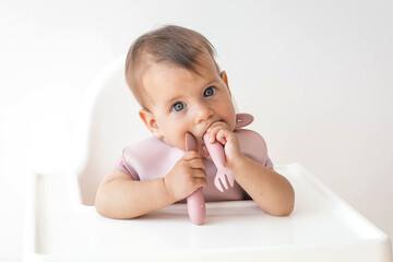 close-up portrait of a cute baby girl infant 8 months old, sitting on a highchair in a baby apron with a fork and spoon, the concept of baby food and baby food.