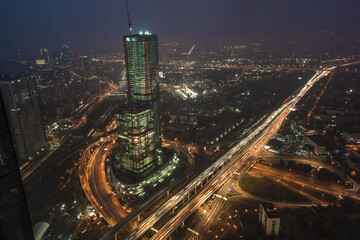 Naklejka premium A vibrant city skyline features a skyscraper in the process of construction, surrounded by busy highways and illuminated streets at night, showcasing the urban life and activity