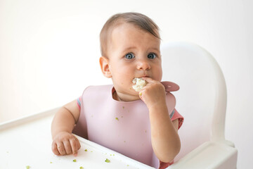 baby's first feeding eating cauliflower food by himself on baby high chair and making mess, close...
