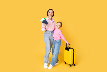 A mother and her preteen daughter stand together with smiles, ready for their travel adventure. The mom holds travel documents, while the girl stands next to a bright yellow suitcase.