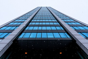 The sight of snow falling on the tall glass facade of a building.