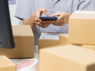 Asian female small business owner, using mobile phone to scan barcode on parcel, preparing for shipment. Asian woman shop owner checking parcel box, working in warehouse office. online store concept.