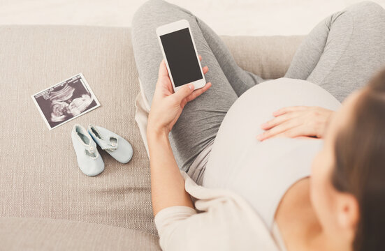 Unrecognizable pregnant woman using smartphone with blank screen, with tiny shoes and sonogram. Young expectant lady surfing net, reading about baby care, buying goods for newborn, copy space