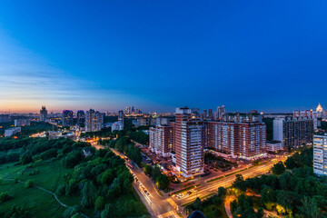 Obraz premium Urban landscape showcases high-rise buildings transitioning into night with city lights twinkling. A large green park contrasts the vibrant skyline, creating a peaceful atmosphere.