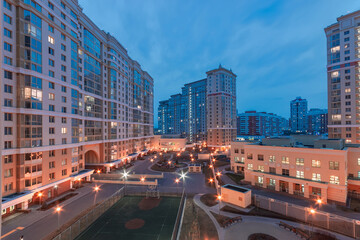 Fototapeta premium The scene captures a modern residential area at twilight, showcasing illuminated apartments and a recreational space with a sports court. The urban landscape is lively and welcoming.