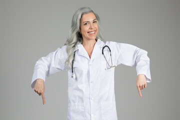 A middle aged woman in a doctor's coat smiles confidently while pointing downwards.