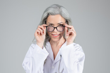 A middle aged woman with long grey hair wears a lab coat and adjusts her glasses with a warm smile. She represents a professional healthcare provider ready to assist patients.