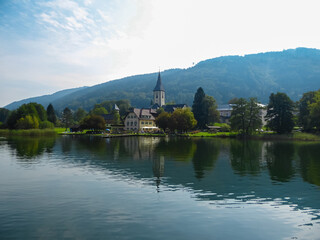 Fototapeta premium Tranquil lakeside scene with Ossiach Abbey nestled amidst rolling hills of Ossiacher Tauern in Carinhtia, Austria. Panoramic boat tour on Lake Ossiach in summer. Benedictine monastery in Austrian Alps
