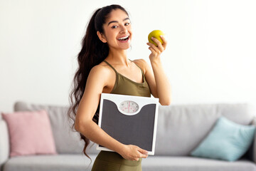 Portrait of millennial Indian lady holding scales and eating apple, choosing healthy diet, panorama. Young Hindu woman preferring wholesome nutrition. Wellbeing and weight loss concept