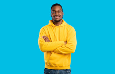 Handsome Black Millennial Man Crossing Hands Posing Smiling To Camera Standing In Studio On Blue Background, Wearing Yellow Hoodie. Shot Of Confident African American Guy