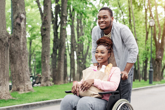 Cheerful young black woman in wheelchair carrying bag with products, going shopping with her boyfriend outdoors, copy space. Happy African American couple returning home from market, walking at park - Powered by Adobe