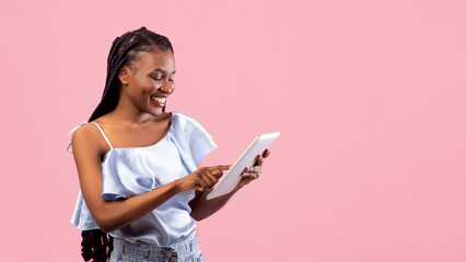 Online life. Pretty young Afro lady using tablet pc for remote work or education on pink studio background. Cool black woman having web conference on touch pad, communicating on internet