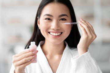 Pretty asian lady applying eye serum and smiling at camera, wearing white bathrobe, holding bottle...