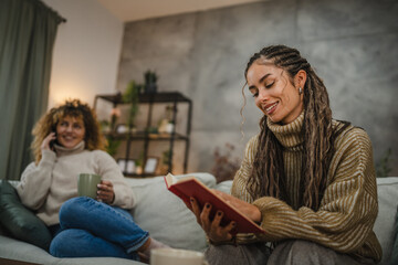 Obraz premium female friends enjoy on sofa one read a book while other use cellphone