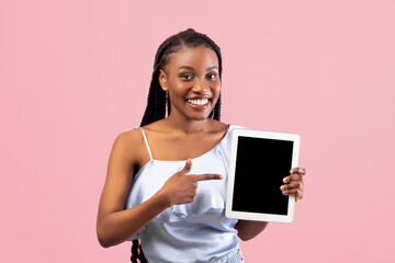 Positive young Afro woman pointing at tablet computer with empty screen on pink studio background, mockup for website or app. Cool black lady presenting touch pad with space for ad