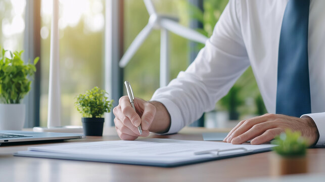 A business professional stamps a document at a desk, symbolizing official certifications or approvals. The presence of a wind turbine and small plants in the background highlights