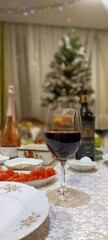 a large glass goblet on a thin stem, half filled with red wine, stands on a New Year's table with a decorated New Year's tree, bottles of sparkling and wine on a blurred background