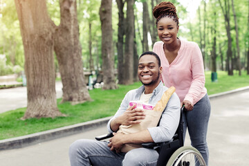 Happy young black guy in wheelchair carrying paper bag with products, going shopping with his girlfriend, outdoors. Millennial African American couple returning home from store, walking at park