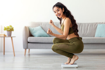 Happy Indian woman sitting on scales, gesturing YES, satisfied with her weight loss result at home,...