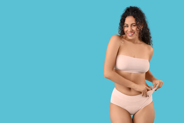 Fototapeta premium Beautiful young happy African-American woman in elegant pink underwear on blue background