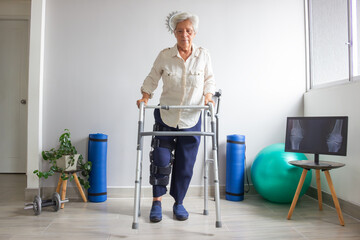 Elderly woman using a walker for support, wearing a leg immobilizer. The setting includes therapy equipment such as a plant, dumbbells, mats, a therapy ball, and a screen displaying a knee X-ray.