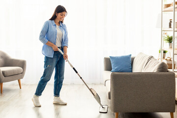 Fototapeta premium Full body length image of smiling young woman in casual clothes washing heated wooden laminate warm floor using microfiber wet mop pad, doing homework cleaning routine. Housekeeping Job Concept