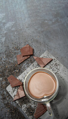 Glass cup of hot chocolate in heart shape on table surrounded by chocolate pieces and black chia seeds on grey textured background, Desert concept in color of the year 2025, mocha mousse, copy space 