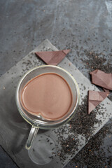 Glass cup of hot chocolate in heart shape on table surrounded by chocolate pieces and black chia seeds on grey textured background, Desert concept in color of the year 2025, mocha mousse, copy space 