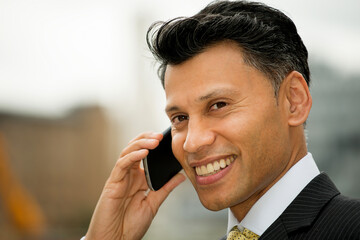 Smiling smartly dressed Asian / Middle eastern businessman holding a mobile phone to his ear, in an outdoor city location.