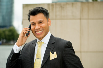 Smiling smartly dressed Asian / Middle eastern businessman holding a mobile phone to his ear, in an outdoor city location.
