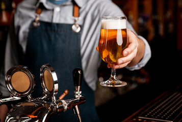 Beer friday evening. Barman in apron gives glass of light beer with foam in dark interior of pub, free space, cropped