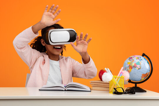 Virtual Reality Concept. Portrait of excited African American girl in VR headset touching air at yellow studio, copy space. Positive teen exploring artificial computer world sitting at desk