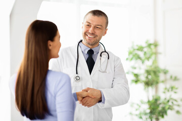 Professional Help. Portrait of smiling middle aged general practitioner shaking hands with woman standing at clinic or hospital. Patient greeting her doctor. Healthcare and Medicine Concept