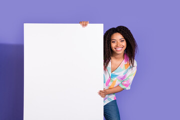 Smiling young woman with black curls standing against a vibrant purple background, holding a blank...