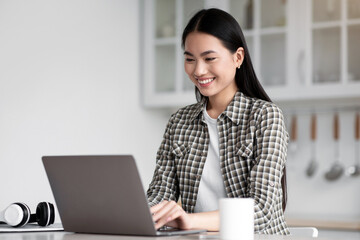 Positive asian lady freelancer sitting at kicthen, working online, using modern laptop, typing on keyboard, sending emails or chatting with clients. Young woman student doing homework, copy space
