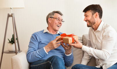 Mature Son Congratulating Elderly Father Giving Him Birthday Gift Sitting On Sofa At Home....