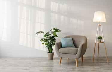 Retro armchair with pillow, glowing lamp, plant in pot on floor on gray wall background, light from window in living room. Blog about interiors, stylish minimalist design, real estate sales and rent