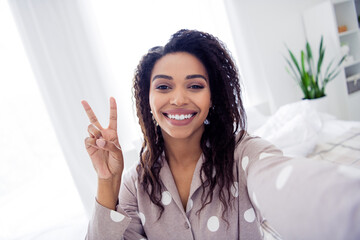 Young woman in polka dot pajama making peace sign and smiling in bright bedroom with natural light