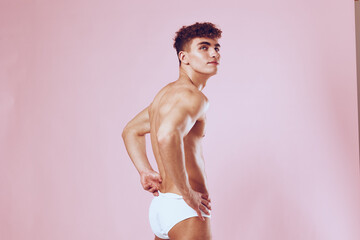 Confident young man posing in white underwear, standing against a pastel pink background, showcasing a fit physique. Body positivity and fashion concept.