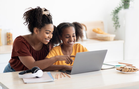 Creative black mother and daughter working on school project, using modern laptop, taking notes, cheerful afro-american woman helping her teen kid with homework, kitchen table, copy space