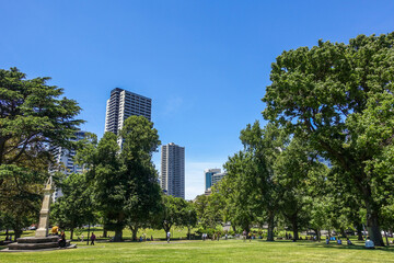 Beautiful environment with lush green lawns, tall trees, and modern skyscrapers in the background at Flagstaff Gardens in Melbourne, Australia. Visitors enjoying the community open space at urban park