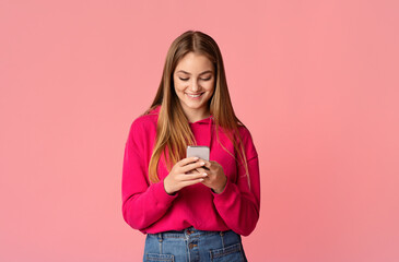 Fototapeta premium Good news in message. Smiling teenage girl checking smartphone isolated on pink background, studio shot
