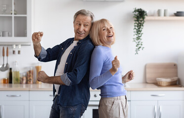 Joyful elderly spouses dancing together at kitchen, happy attractive senior man and woman in casual standing back to back and making dance moves, enjoying weekend at home, copy space