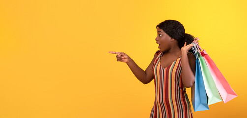 Big sales concept. Shocked young black woman with colorful shopping bags pointing at blank space over orange studio background, panorama. Surprised African American lady cannot believe huge discounts