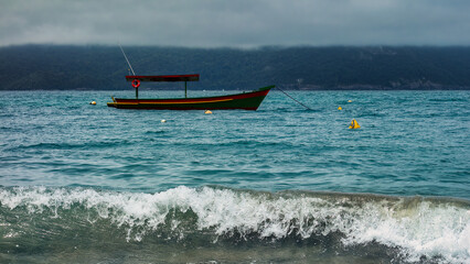Detail of small fishing boat in blue sea, with wave breaking on the beach, on cloudy day
