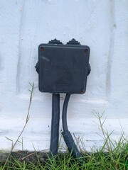 A close-up of a black electrical junction box mounted on a white wall. Two black cables enter the box from below