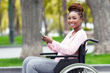 Happy disabled black woman in wheelchair using cellphone, checking social media, browsing web at...