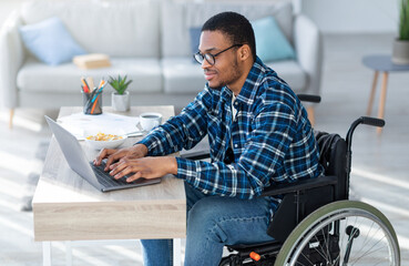 Online work for disabled people. Handicapped black guy in wheelchair using laptop for remote job at...